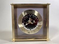 Front view of square brass mantle clock with world map and time zones around dial, airplane second hand visible.
