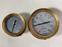 Top-down photo showing two brass pressure gauges of different sizes side by side, one with white dial and one with gray dial, both with brass casing.