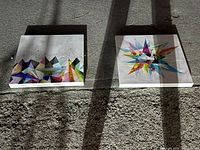 Photo of two square paintings with colorful geometric designs on white surfaces taken outdoors on concrete ground showing possible natural light shadows.