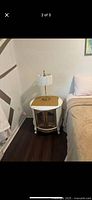 White painted drum-style nightstand positioned beside a bed with a white lamp on top. Same style as first, showing matching pair. Glass doors and shelf visible.