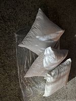 Three medium size white feather pillows arranged on a dark carpeted floor with a transparent plastic sheet beneath them.