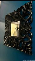 Front view of the square black uniquely designed decorative mirror showing intricate carved scroll and leaf patterns on the thick frame.