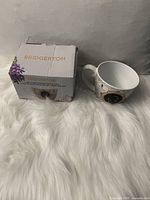 Photo of Bridgerton-branded teacup next to its original box on white faux fur surface, showing front and side of box and cup handle.