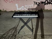 Casio keyboard displayed on a foldable X-style metal stand outdoors on a rough pavement with graffiti wall background.