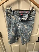 Front view of light blue denim kids shorts with unicorn and star print, hung on a hanger showing details like pockets and button fly.