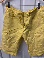 Front view of bright yellow men's shorts hanging on a hanger, showing button and zipper closure, front pockets, and fabric texture.