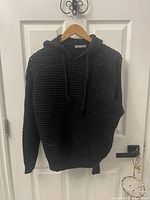 Front view of black knitted sweater hoodie hung on wooden hanger against a white door. The sweater has an open weave pattern and drawstrings on the hood.