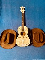 Full view of the 1966 Emenee plastic Western folk guitar centered between two brown cowboy hats on a blue background.