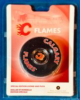 Front view of sealed packaging showing black Calgary Flames hockey puck with embedded loonien coin in center, red and white graphics with team and mint branding.