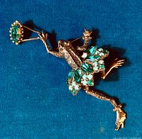 Top view of the frog brooch showing the green rhinestones on the tutu and wand against a blue background.