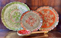 Three round scalloped edge trays with Florentine gesso painted details in various colors, arranged vertically against wood background with a wooden decorative spoon in front