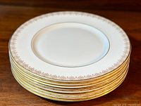 Stack of 8 white dinner plates with a delicate gold lace pattern around the rim and a thin gold band near the center, showing decorative pattern and edge design.