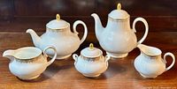 Full set displayed on wooden table, showing tea pot, coffee pot, milk jug, creamer, and sugar bowl with lids and handles, in white fine bone china with gold lace detail.