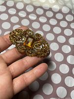 Front view of brooch displaying central rectangular light brown-yellow gemstone surrounded by smaller stones in ornate gold-tone filigree setting, held in hand.