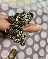 Close-up of butterfly ring held between fingers showing detailed filigree with embedded clear round gemstones.