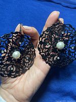 Close view of both large black filigree round earrings with pearl centerpiece held in hand to highlight size, design details and rhinestone accents.