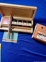 Opened wooden box showing four jars of acrylic artists ink, two calligraphy pens, packet of nibs, and instruction card lying on blue cloth surface.