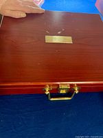 Closed wooden rectangular case with brass handle and latch, displaying small brass plaque on top.