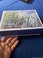 Decorative box with artwork and title 'Irises by Monet' held in hand against blue background.