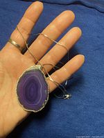 Photo of hand holding necklace with large violet agate slice pendant on silver chain, showing front view of pendant.