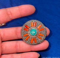 Front view of the round brooch showing turquoise central stone and orange stones on textured silver-tone base held by hand.
