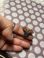 Close-up of the ring held in hand showing the side profile with blue-green stones on the band and red stones surrounding the central yellow stone.