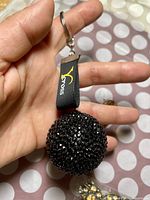 Hand holding a round black keychain ball encrusted with small sparkling stones, showing the Cross logo on the attached black fabric loop.