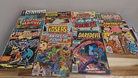 Lot of 12 vintage comic books on wood surface showing titles including Captain Marvel, Daredevil, Son of Dracula, Hercules, Fantastic Four, Losers, William Tell, and Shogun.