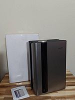 Front view of the silver and black dehumidifier standing on a wooden surface next to its white box and instruction manual.