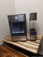 Front angled view of a Profile stainless steel countertop ice maker with a clear plastic front panel and adjacent tall detachable water reservoir on a wooden table.