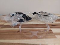 Top view of four pairs of clear safety glasses on wooden surface, two pairs remain in sealed packaging, two pairs out of packaging showing translucent frames and wrap-around lenses.