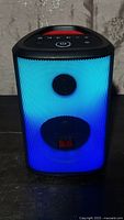 Front view of the black Monster Sparkle speaker illuminated with bright blue LED lights from the front grille, showing the digital touch control panel on top and Monster logo on front.