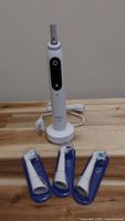 Electric toothbrush handle with power button, charging base behind, 3 replacement brush heads in front with blue covers.