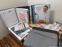 Photo showing a Samsung gray tablet cover with a front zippered pocket, colorful ring-bound planner with motivational pages, notebook, and various planner stickers next to a cookbook.