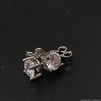 Pair of sterling silver diamond stud earrings shown on black background with one earring laying flat and the other at an angle highlighting the stone and prong setting.