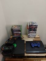 Wide view showing original Xbox with two wired controllers, PS3 Slim with blue controller, and stacks of game cases.
