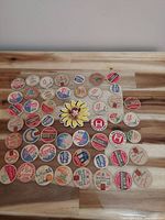 Top-down photo showing all 51 vintage milk bottle caps arranged in rows on a wooden surface revealing various colors, text, logos, and one sunflower design.