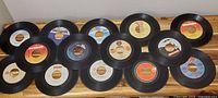 Top-down view of 15 assorted 7-inch 45 RPM vinyl records arranged in 3 rows on a wooden surface, showing various colored record labels and labels for artist and song information.