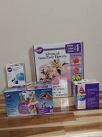Photo displaying all four Wilton cake decorating products arranged on a wooden surface: Advanced Gum Paste Flowers kit, Icing Colours, Airbrush Set, and Color Right Colour System.