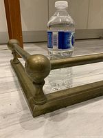 Close-up image of one corner of the brass fireplace fender showing spherical finial post, horizontal bars, and original patina. A water bottle is placed inside for scale.