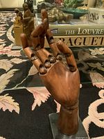 The carved jointed wooden hand is posed upright on a patterned carpet, showing the fingers curled in a natural gesture. Background includes decorative objects and books.