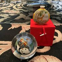 Clear glass orb featuring two pandas with bamboo and flowers inside, sitting on a surface with patterned fabric background alongside a red box with miniature painted glass bottle on top.