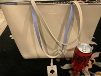 Front view of large cream-colored Kate Spade tote bag with gold logo and spade-shaped hanging tag, placed next to a soda can for size reference.