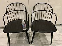 Front view of two black chairs with rounded metal backrest and wooden seats. One bottle on seat showing scale.