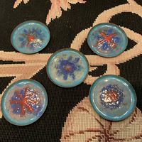 Five small round copper and enamel plates/coasters laid out in a group showing varied blue and red starburst or floral abstract patterns, on a dark patterned fabric surface.