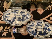 Two blue and white ceramic trays with Asian-themed landscape patterns showing pagodas, trees, and mountains. Trays have scalloped edges and are displayed on a floral patterned surface with a water bottle for scale.