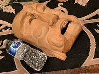 Side angle of hand-carved wooden Asian face mask alongside bottled water to illustrate size and detail.