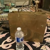 Front view of brass magazine rack shaped like a shopping bag with twisted handles. Shows size relative to water bottle and visible patina and surface wear.