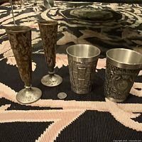 Four vintage etched metal items arranged on a dark and patterned surface, showing two tall vases with floral etching and two shorter cups with detailed embossing.