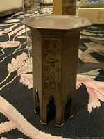 Small vintage brass carved table/stand with octagonal top and carved panels showing figures and designs, standing on a patterned carpet.